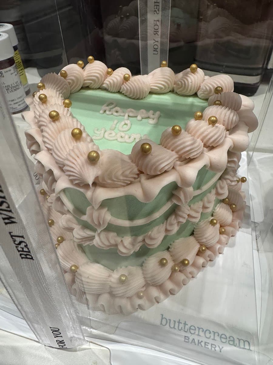 A multi-tiered white wedding cake adorned with pearls and topped with a figurine couple, displayed on a wooden table alongside cupcakes.