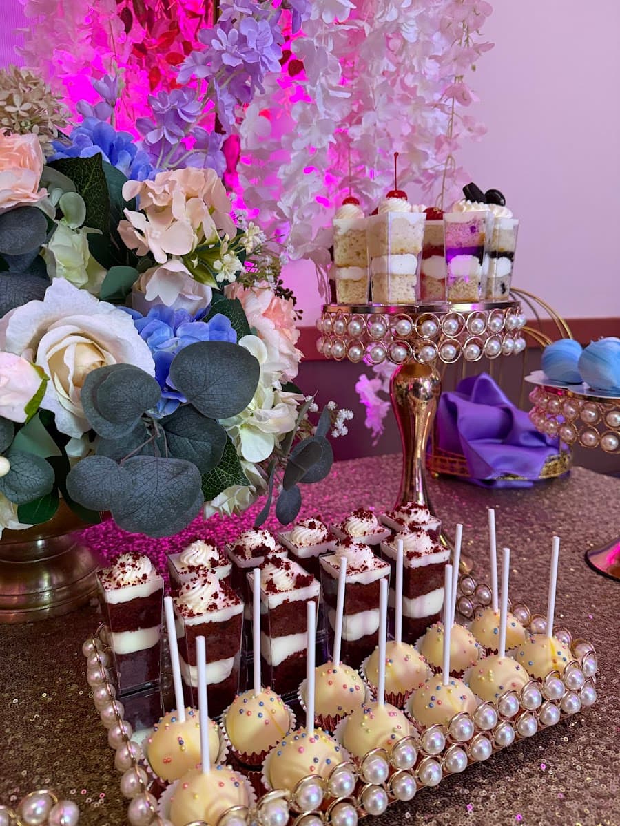 A multi-tiered white wedding cake adorned with pearls and topped with a figurine couple, displayed on a wooden table alongside cupcakes.