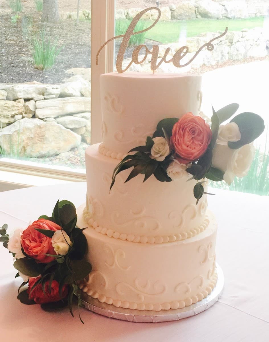 A multi-tiered white wedding cake adorned with pearls and topped with a figurine couple, displayed on a wooden table alongside cupcakes.